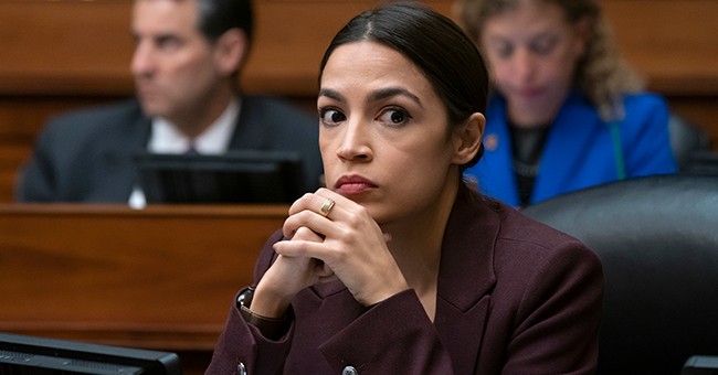 AOC sits in the House with hands clasped and a determined stare as the mercifully blurred image of Debbie Wasserman-Schultz looms in the background.